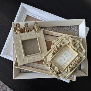 May include: A selection of decorative picture frames in cream and white. The frames vary in shape and size, with ornate detailing and embellishments. Some frames feature small angel figurines. One frame has the word "FIGI" printed on it.