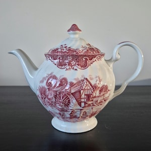 May include: A white ceramic teapot featuring a red transferware pattern depicting a watermill scene. The teapot has a curved handle, a spout, and a matching lid with a finial. The body of the teapot has a subtle swirl texture and a wide base.