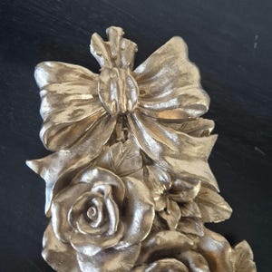 May include: A gold-coloured decorative wall ornament featuring a sculpted bow at the top with long, flowing ribbons cascading down. Below the bow are detailed, three-dimensional roses and leaves, all finished in a metallic gold tone with highlights.