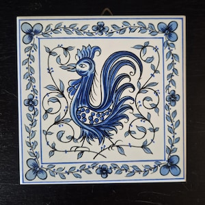 May include: A square ceramic tile featuring a hand-painted blue cockerel centred within a decorative border of blue flowers and vines. The design is rendered in shades of blue on a white background, with a metal hanging loop attached at the top.