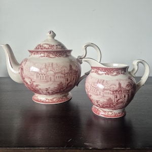 May include: A ceramic teapot and matching creamer set featuring a cream-coloured base with a detailed maroon transferware pattern depicting architectural buildings and figures. Both pieces have a pedestal base, curved handles, and ornate trim.