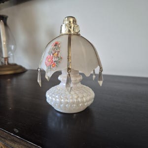 May include: A small table lamp with a white hobnail glass base and a frosted glass shade. The shade features a floral decal with pink and orange flowers, metallic gold-tone metal accents, and hanging clear glass crystal-style teardrop prisms.