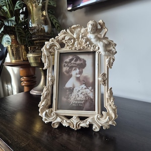 May include: An off-white, ornate picture frame featuring intricate scrollwork and a sculpted cherub figure at the top right corner. The frame holds a sepia-toned photograph of a person holding flowers, with the text "photo frame 10cm x 15cm" visible.