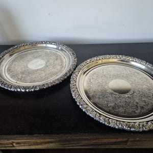 May include: Two silver oval serving trays with detailed floral patterns and decorative edges. The trays are on a dark surface, highlighting their reflective surfaces. The trays are likely vintage or antique.