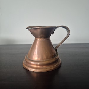 May include: A small copper jug with a wide, flared rim, a rounded body, and a curved handle. The metal surface has a polished finish with subtle variations in tone, featuring a distinct tiered base and a small pouring spout at the top.