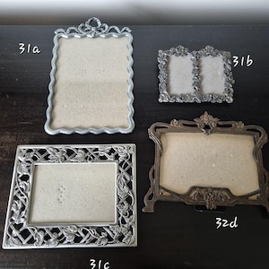 May include: Four metal picture frames labelled 31a, 31b, 31c, and 32d. Frame 31a is a tall silver rectangle with a bow top. Frame 31b is a double silver frame with floral borders. Frame 31c is a silver rectangle with lattice and leaf details. Frame 32d is a bronze-toned frame with ornate scrollwork.