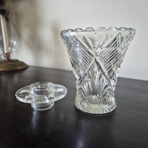 May include: A clear glass vase with a flared, scalloped rim and a geometric diamond-cut pattern on the exterior. Beside the vase sits a separate, circular glass flower frog insert featuring a central opening and five smaller circular holes.