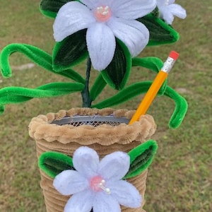 May include: A handmade craft flower arrangement in a brown pot. The arrangement features white flowers with pink centers, green leaves, and green stems. A yellow pencil with a red eraser is placed in the pot.