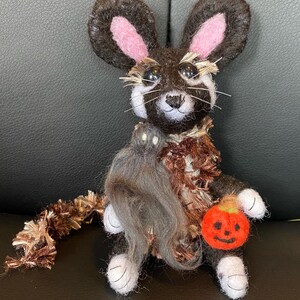 May include: A brown and black felt mouse wearing a brown and white costume with a ghost and a pumpkin. The mouse has pink ears and black eyes. The mouse is holding a small orange pumpkin with a jack-o-lantern face.