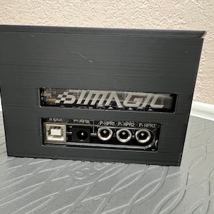 Puede incluir: Caja negra con el texto "SIMAGIC" en la parte delantera. La caja tiene un puerto USB, un puerto de alimentación y otros tres puertos etiquetados como "P-HPRI", "P-HPR2" y "P-HPR3".