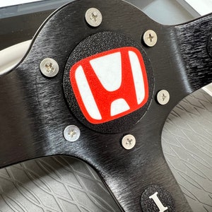 May include: Close-up of a black steering wheel with a red and white Honda logo in the centre. The steering wheel has a brushed aluminium finish and silver screws.