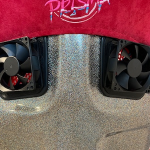 May include: Close-up of a black gaming chair with two black fans on the back. The chair is covered in a silver glitter fabric. The top of the chair is red with the word "PRISMA" embroidered in pink and blue.