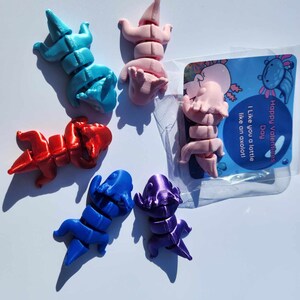 May include: Five small, colorful, articulated axolotl figurines. The axolotls are in various poses and are made of a smooth, plastic-like material. The axolotls are blue, red, pink, purple, and light blue. The background is white. The image also includes a small card with the text "Happy Valentine's Day. I like you a lot, like an axolotl."