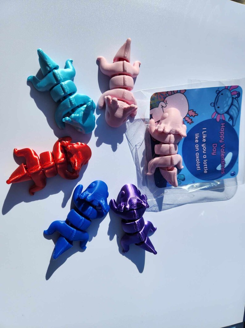 May include: Five small, colorful, articulated axolotl figurines. The axolotls are in various poses and are made of a smooth, plastic-like material. The axolotls are blue, red, pink, purple, and light blue. The background is white. The image also includes a small card with the text "Happy Valentine's Day. I like you a lot, like an axolotl."