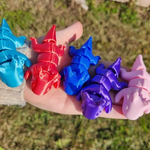 May include: Five 3D printed figurines of small, segmented creatures in various colors. The figurines are blue, red, purple, and pink. They are arranged in a row on a hand.