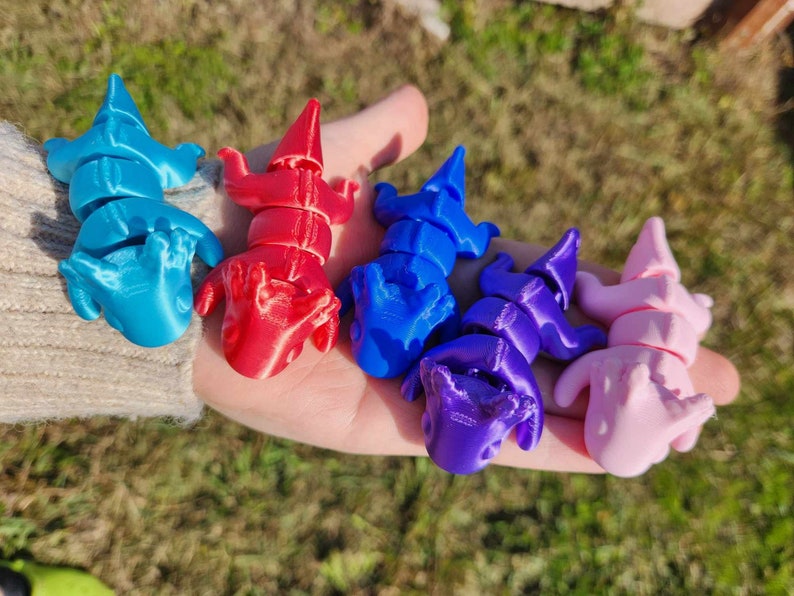 May include: Five 3D printed figurines of small, segmented creatures in various colors. The figurines are blue, red, purple, and pink. They are arranged in a row on a hand.