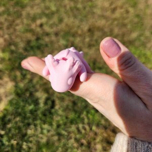 May include: A pink, 3D printed, small, cartoon-like creature with a red heart on its chest. The creature is being held in a person's hand.