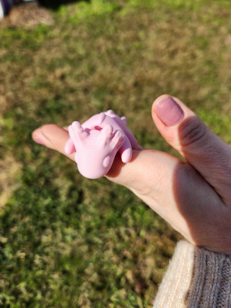 May include: A pink, 3D printed, small, cartoon-like creature with a red heart on its chest. The creature is being held in a person's hand.