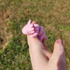 May include: A pink, 3D printed, curled up, cartoon-like creature with a pointed head and a tail. The creature is being held in a person's hand.