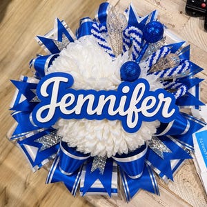 May include: A decorative homecoming mum featuring a central white chrysanthemum flower, layered with royal blue and silver glitter ribbons. The name Jennifer is displayed in white script lettering on a blue background, accented by blue glitter spheres.