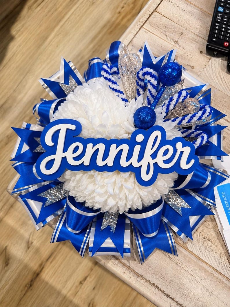 May include: A decorative homecoming mum featuring a central white chrysanthemum flower, layered with royal blue and silver glitter ribbons. The name Jennifer is displayed in white script lettering on a blue background, accented by blue glitter spheres.