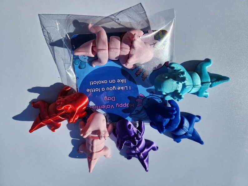 May include: A set of six colorful axolotl figurines in a clear plastic bag. The figurines are in various poses and are made of a smooth, glossy material. The bag also contains a Valentine's Day card with the text "Happy Valentine's Day. I like you a lottie like an axolotl."