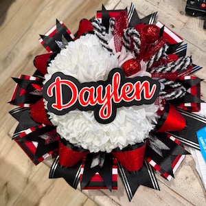 May include: A decorative homecoming mum featuring a large white chrysanthemum centre with a black and red nameplate reading Daylen. The design includes layered ribbons in black, red, and white, accented with glittery red spheres and black and white striped mesh.