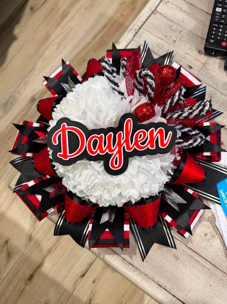 May include: A decorative homecoming mum featuring a large white chrysanthemum centre with a black and red nameplate reading Daylen. The design includes layered ribbons in black, red, and white, accented with glittery red spheres and black and white striped mesh.