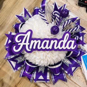 May include: A decorative homecoming mum featuring a central white chrysanthemum surrounded by layers of purple and white satin ribbons, silver glitter accents, and two purple glitter spheres. The name Amanda is displayed in white script.