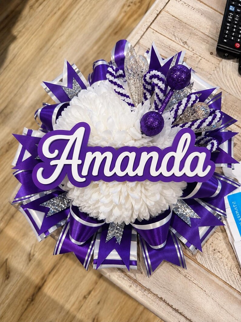 May include: A decorative homecoming mum featuring a central white chrysanthemum surrounded by layers of purple and white satin ribbons, silver glitter accents, and two purple glitter spheres. The name Amanda is displayed in white script.