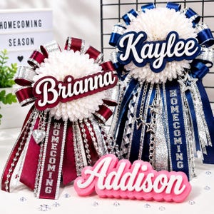 May include: Two large homecoming mums with white chrysanthemum centres, layered with maroon and navy blue satin ribbons, silver glitter accents, and hanging chains. The names Brianna and Kaylee are displayed in script, with a pink Addison nameplate.