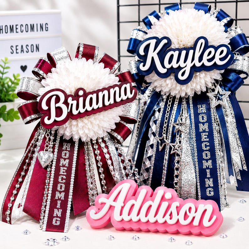 May include: Two large homecoming mums with white chrysanthemum centres, layered with maroon and navy blue satin ribbons, silver glitter accents, and hanging chains. The names Brianna and Kaylee are displayed in script, with a pink Addison nameplate.