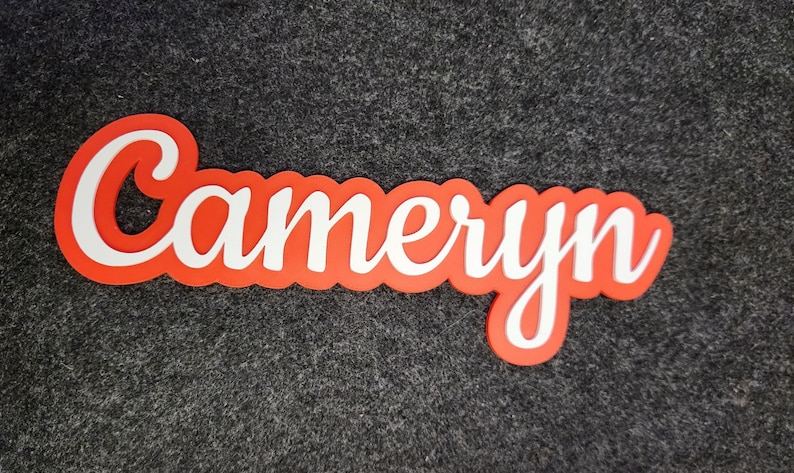 May include: A custom name sign featuring the name Cameryn in a white cursive script font. The lettering is layered over a solid orange background that follows the contour of the letters, creating a bold, high-contrast decorative name plaque.