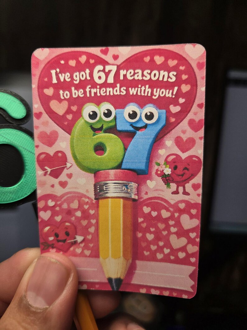May include: A Valentine's Day card with the text "I've got 67 reasons to be friends with you!" features a pencil with a pink eraser and the numbers 6 and 7 with cartoon eyes. The background is pink with hearts.