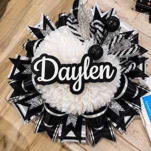 May include: A decorative homecoming mum featuring a large white chrysanthemum centre with the name Daylen in black script lettering. The design includes layers of black and white satin ribbons, silver glitter accents, and black glitter spheres.