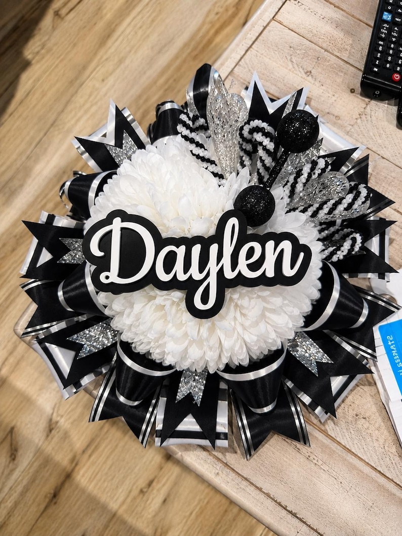 May include: A decorative homecoming mum featuring a large white chrysanthemum centre with the name Daylen in black script lettering. The design includes layers of black and white satin ribbons, silver glitter accents, and black glitter spheres.