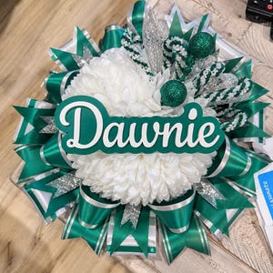May include: A decorative homecoming mum featuring a large white chrysanthemum centre, layered with emerald green and silver satin ribbons, glittered accents, and a prominent green and white cutout sign displaying the name Dawnie in script.