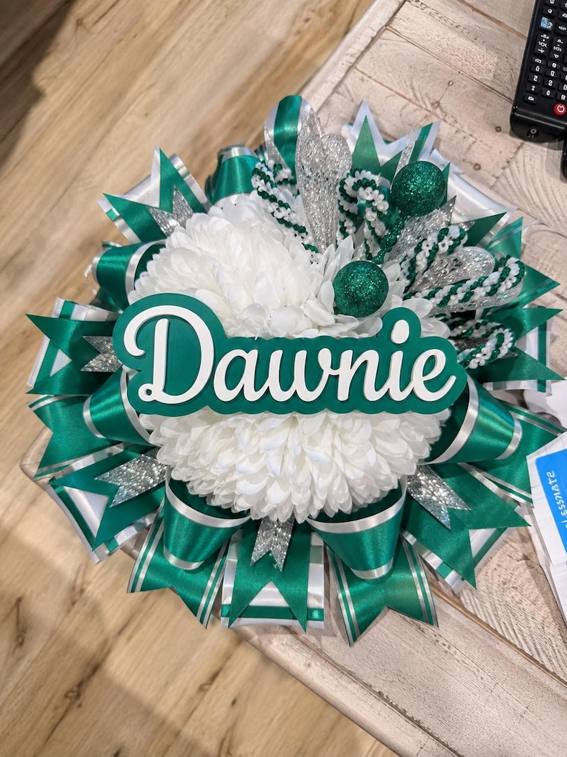 May include: A decorative homecoming mum featuring a large white chrysanthemum centre, layered with emerald green and silver satin ribbons, glittered accents, and a prominent green and white cutout sign displaying the name Dawnie in script.