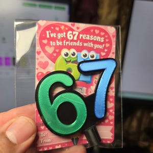 May include: A friendship-themed gift tag with the text "I've got 67 reasons to be friends with you!" The tag features a green and blue number "67" design. The background is pink with heart shapes. The tag is in a clear plastic sleeve.