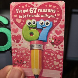 May include: A Valentine's Day card with the text "I've got 67 reasons to be friends with you!" features a pencil with a pink eraser and the numbers 6 and 7 with cartoon eyes. The background is pink with hearts.