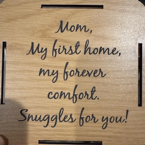 May include: A light-colored wooden coaster with rounded corners and cut-out slots. The coaster features the text "Mom, My first home, my forever comfort. Snuggles for you!" in a black, cursive font.