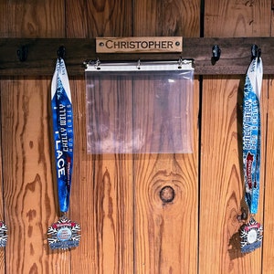 May include: A wooden wall mounted medal rack with the name "CHRISTOPHER" engraved on it. Three medals are hanging on hooks, two blue and one silver. The medals are from the "Chilly Willy Run Series".