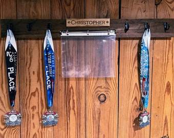 May include: A wooden wall mounted medal rack with the name "CHRISTOPHER" engraved on it. Three medals are hanging on hooks, two blue and one silver. The medals are from the "Chilly Willy Run Series".