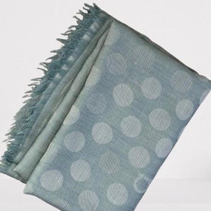 May include: A light blue scarf featuring a repeating pattern of large, pale white polka dots. The fabric has a subtle woven texture and is finished with a frayed, fringed edge along one side, creating a soft and textured appearance throughout.