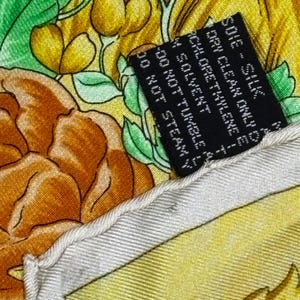 May include: Close-up of a silk scarf with a floral pattern in shades of yellow, orange, and green. A black tag with white text is visible, indicating care instructions. The scarf's edge is a light cream color.