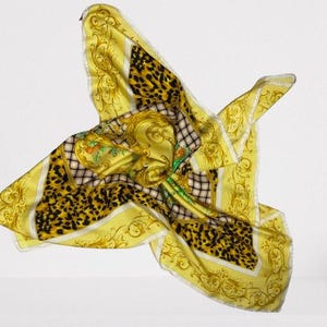 May include: A yellow silk scarf with a leopard print and ornate gold baroque detailing. The scarf features a central design with a portrait and floral accents, framed by a checkered pattern and white borders. The scarf is in motion, appearing to float.