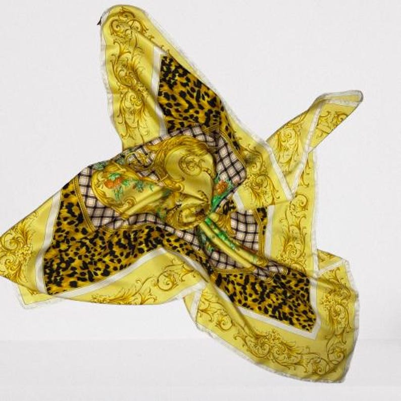 May include: A yellow silk scarf with a leopard print and ornate gold baroque detailing. The scarf features a central design with a portrait and floral accents, framed by a checkered pattern and white borders. The scarf is in motion, appearing to float.