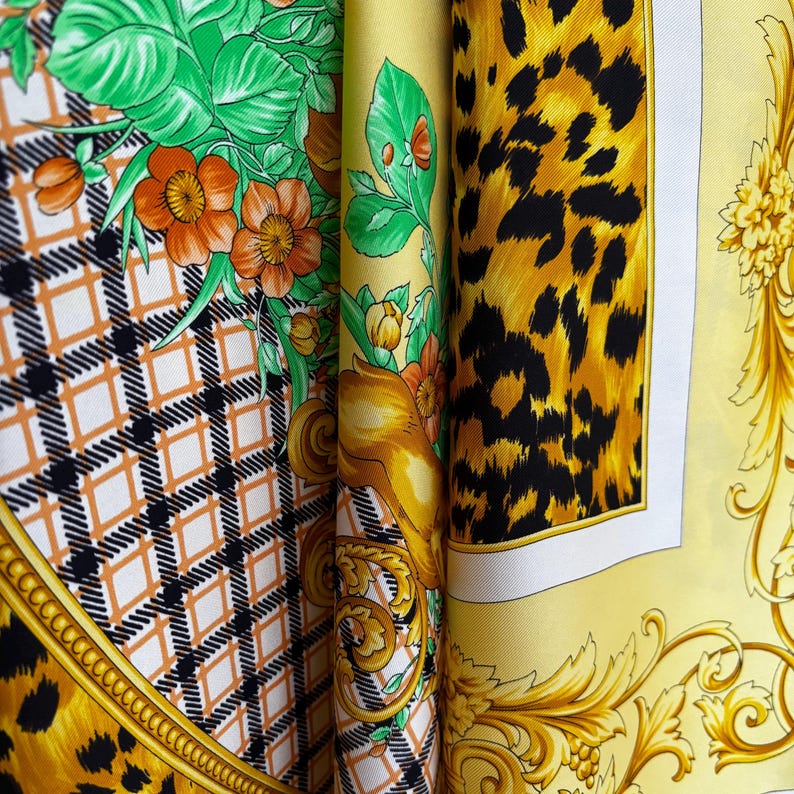 May include: A folded silk scarf with multiple patterns. The scarf features a leopard print, floral designs with orange flowers and green leaves, and a geometric diamond pattern. Gold and yellow tones dominate the design, with black and white accents.