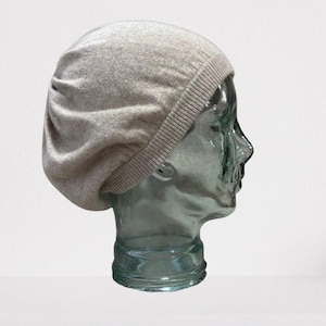 May include: A light gray knit beret hat. The hat is made of a soft, textured material and features a ribbed band at the base. The beret is displayed on a clear glass head form, showcasing its shape and style.
