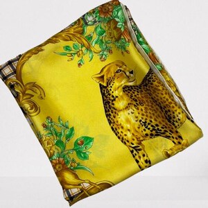 May include: Yellow silk scarf with a cheetah design. The scarf features a detailed illustration of a cheetah with brown spots, set against a yellow background. Floral and gold baroque accents frame the cheetah.
